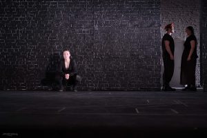 Szene aus einem Theaterstück: Eine Frau hockt an eine Wand gelehnt und blickt in den Zuschauerbereich. Im Hintergrund stehen sich ein Mann und eine Frau gegenüber.
