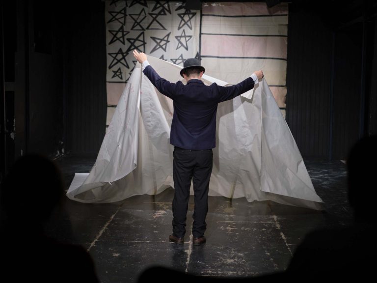Szene aus einem Theaterstück: Ein Mann steht mit dem Rücken zum Publikum und breitet ein weißes Tuch aus.
