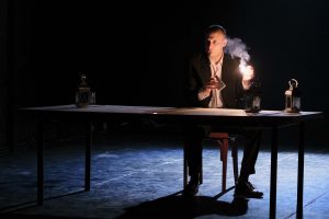 Ein Theaterschauspieler sitzt an einem Schreibtisch in einem dunklen Raum und zündet ein Streichholz an.